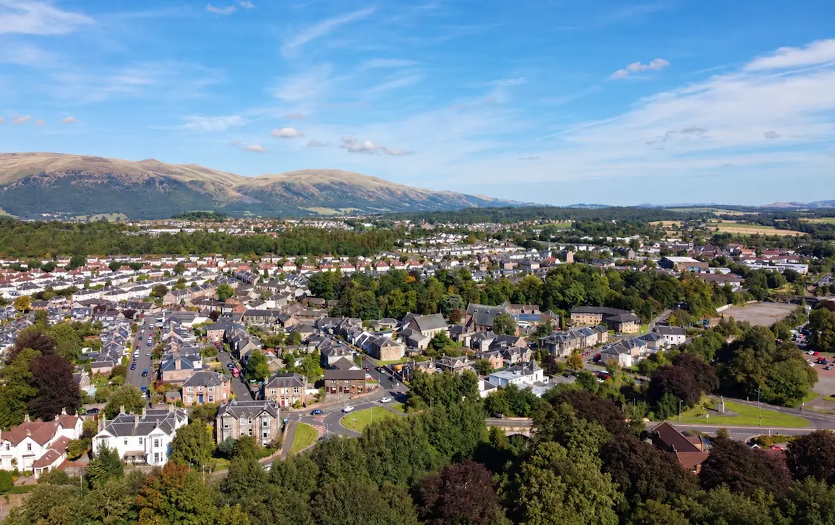 Aerial view of a Clackmannanshire town, with housing, tree-lined streets and green hills beneath a blue sky.