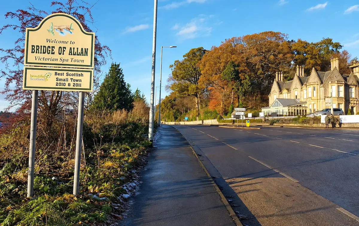 A Bridge of Allan welcome sign stands beside a road, with autumn trees and a stone Victorian building beneath a clear blue sky.