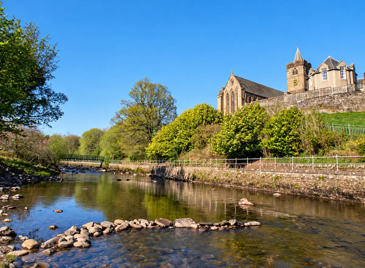 Dunblane Cathedral's stone tower and church rise above a tree-lined river, with rocks along the water beneath a clear blue sky.