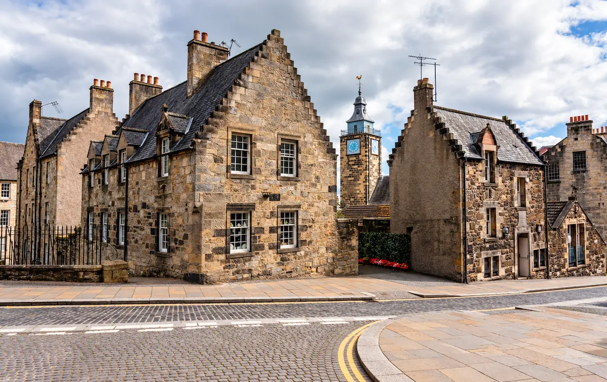 Historic stone buildings with stepped gables and a clock tower line a cobbled street in Stirling beneath a cloudy sky.
