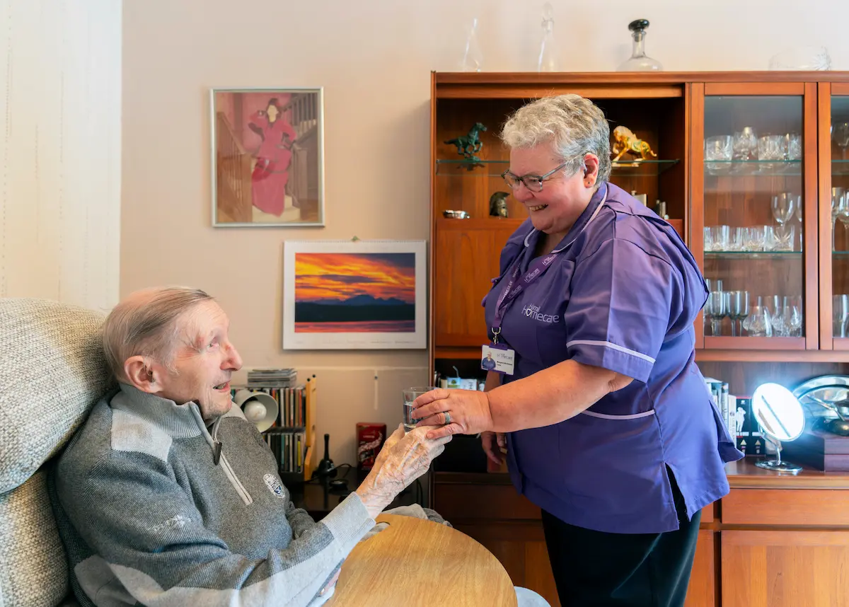 A Hamill Homecare carer handing a glass of water to a client in his living room