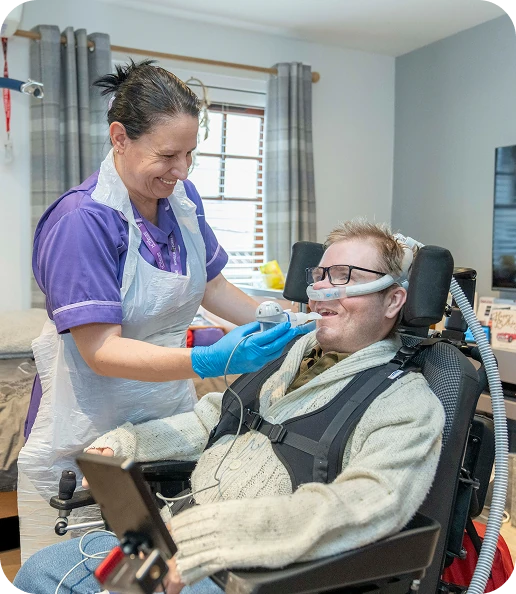 A smiling carer in purple uniform and blue gloves supports a man wearing glasses and a breathing mask in his wheelchair at home.