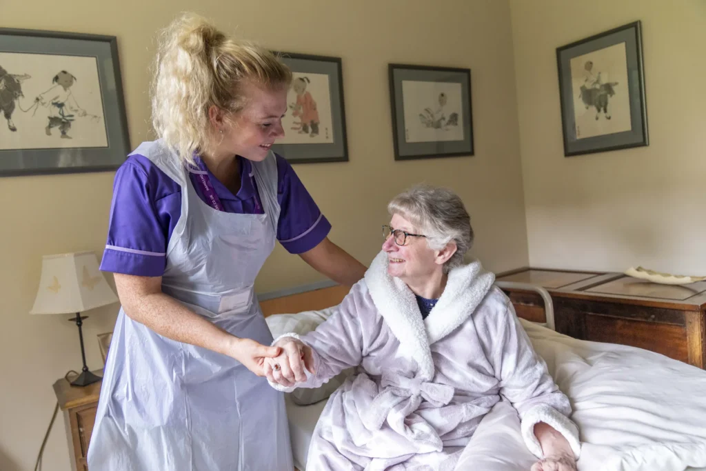 A carer in an apron helps an older woman up from her bed